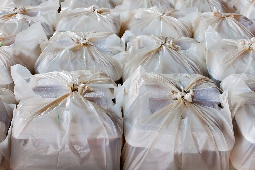A close-up view of multiple tightly packed white plastic rubbish bags, each gathered and sealed with twist ties, arranged on a flat surface likely outdoors or on a driveway. The bags are glossy and semi-transparent, revealing the contents inside which appear to be general waste or refuse, with some hints of grey and black materials. The bags are uniformly positioned, creating a repetitive pattern across the foreground and middle ground, with slight overlapping. The background is out of focus, suggesting a clean, neutral environment suitable for rubbish collection. This scene is representative of waste ready for removal in a typical urban or suburban setting, resonating with services offered by Commercial Waste Removal St Johns Wood or similar rubbish removal providers servicing the local area.
