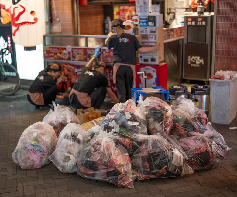 A white outdoor rubbish collection vehicle with an open rear compartment is positioned on a cobblestone street in an urban area, with a worker in a blue uniform and orange high-visibility vest emptyin