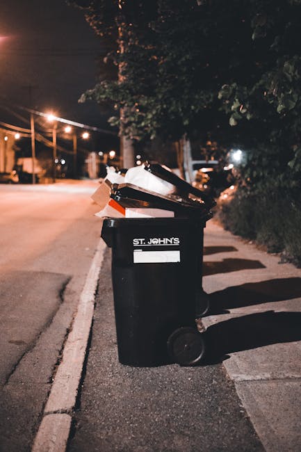 The image depicts a row of black garbage bags filled with waste, neatly arranged on a grassy area in front of a modern multi-storey residential building. The building features large glass windows with